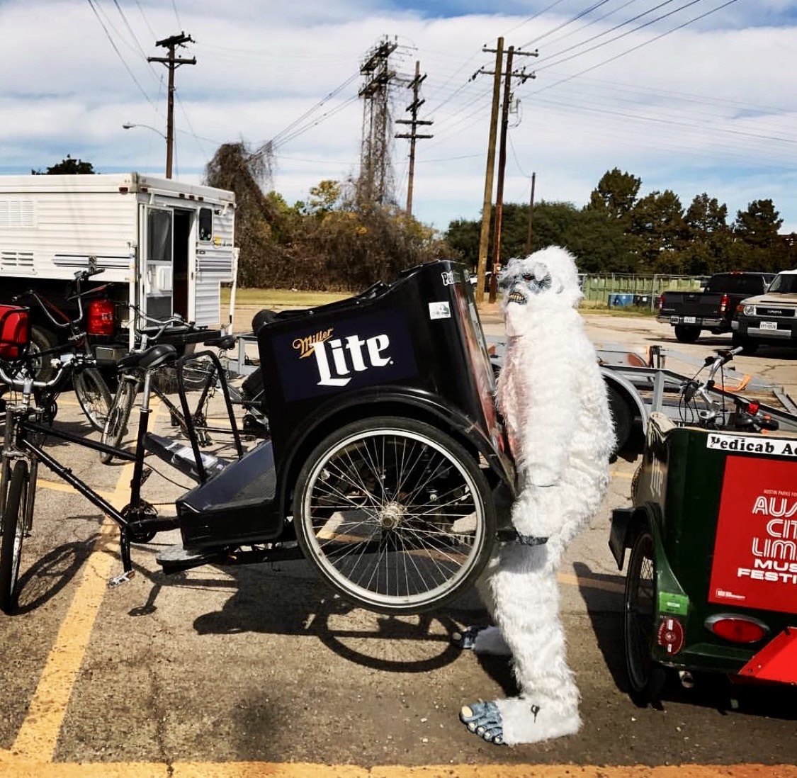 A pedicab driver in a full white yeti costume beside a Miller Lite branded carriage and an Austin City Limits branded pedicab