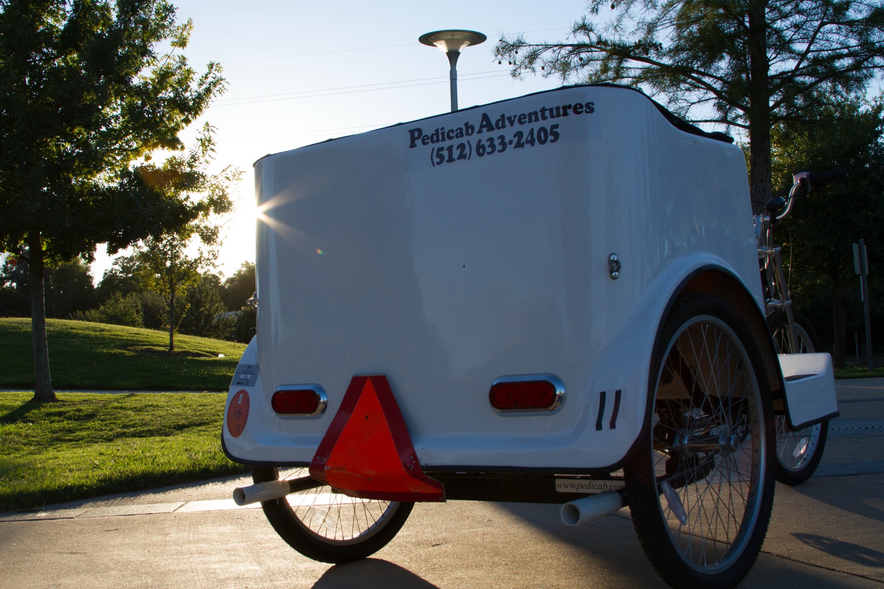 A white Pedicab Adventures carriage lettered with the company name and phone number, backlit by the setting sun
