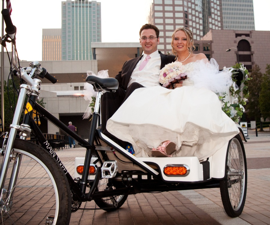 A bride and groom riding in a white pedicab carriage decorated with tulle and flowers, downtown high-rises behind them